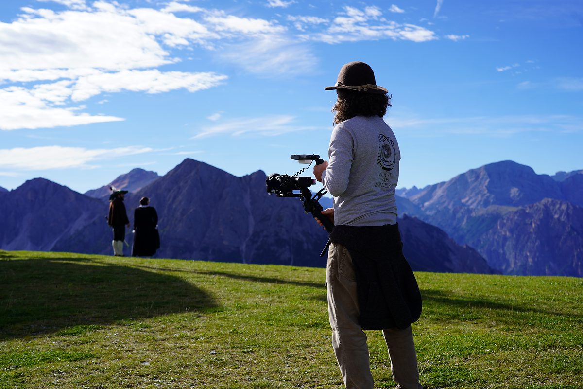 Der Filmemacher steht mit der Kamera auf der Gipfelwiese, vor ihm gehen zwei Menschen in Tracht auf die Berge zu.