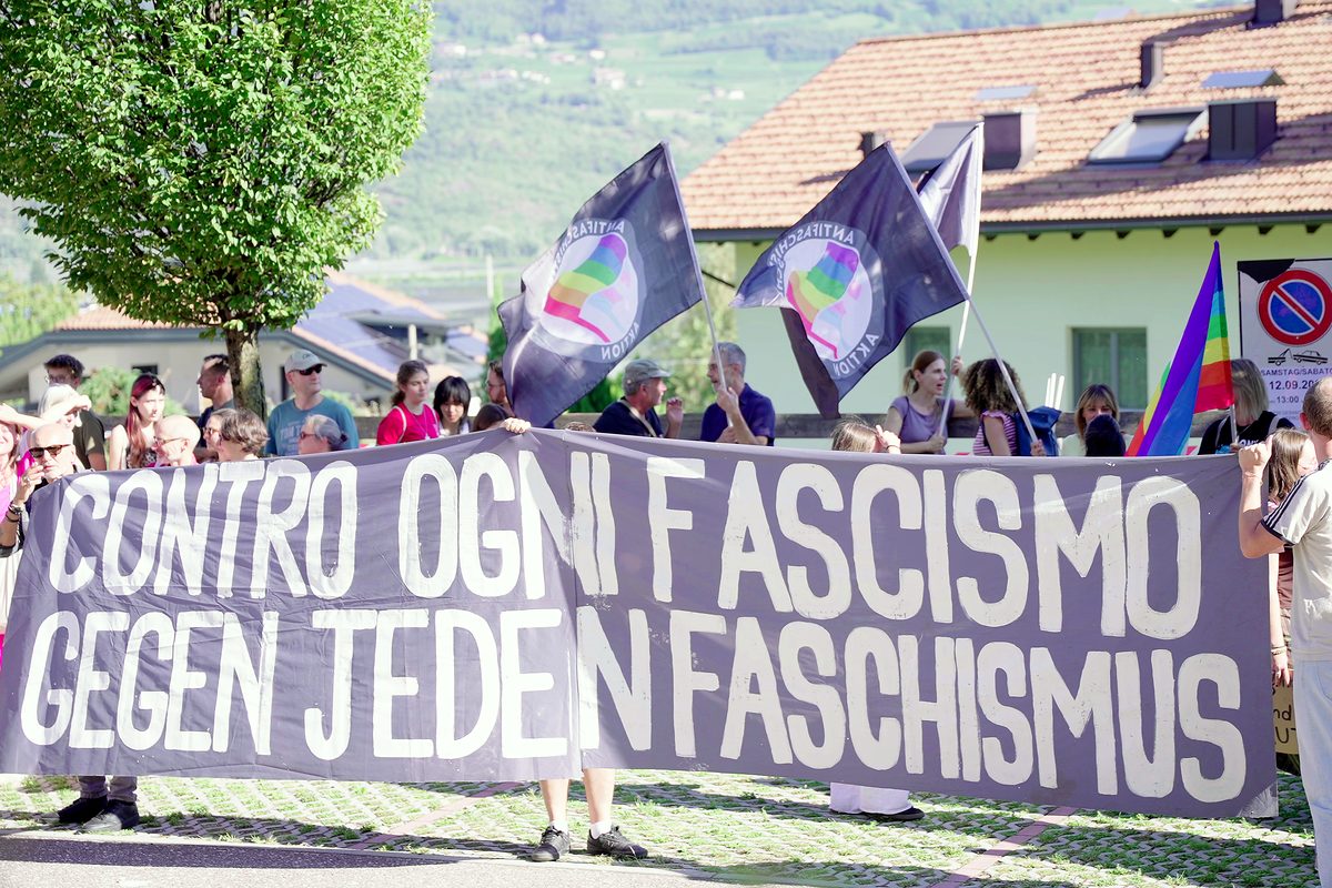 Menschen halten bei einer Kundgebung ein zweisprachiges Transparent, dahinter wehen Fahnen.
