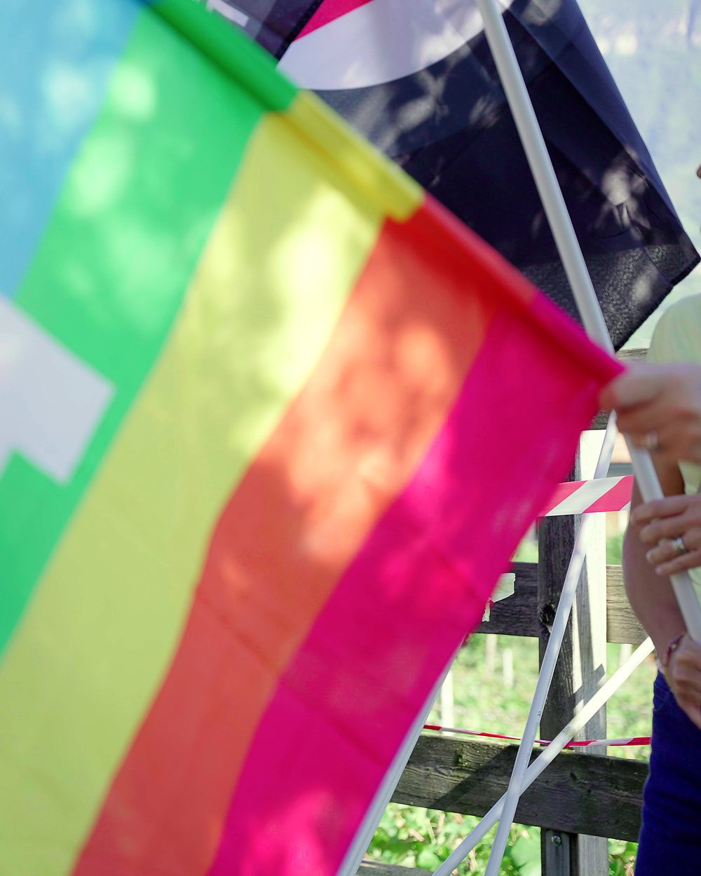 Eine große Regenbogenfahne füllt das Bild, dahinter unscharf die Wartenden.