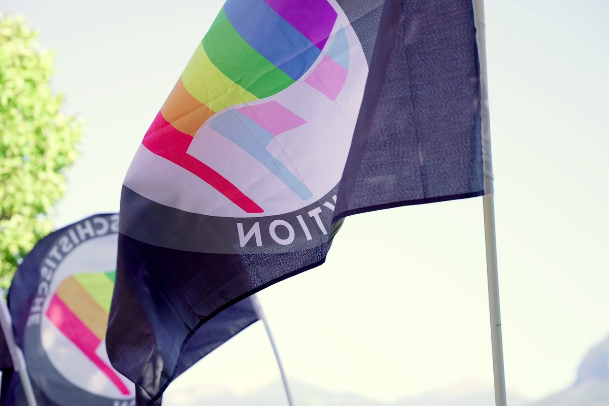 Eine schwarze Fahne mit Regenbogenkreis weht gegen den hellen Himmel.